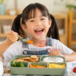 Seorang anak menikmati menu makan siang sehat berisi nasi, ayam, sayuran, telur, dan buah di meja makan dengan suasana ceria. (Foto: dok previews.sriwijayaterkini.id)